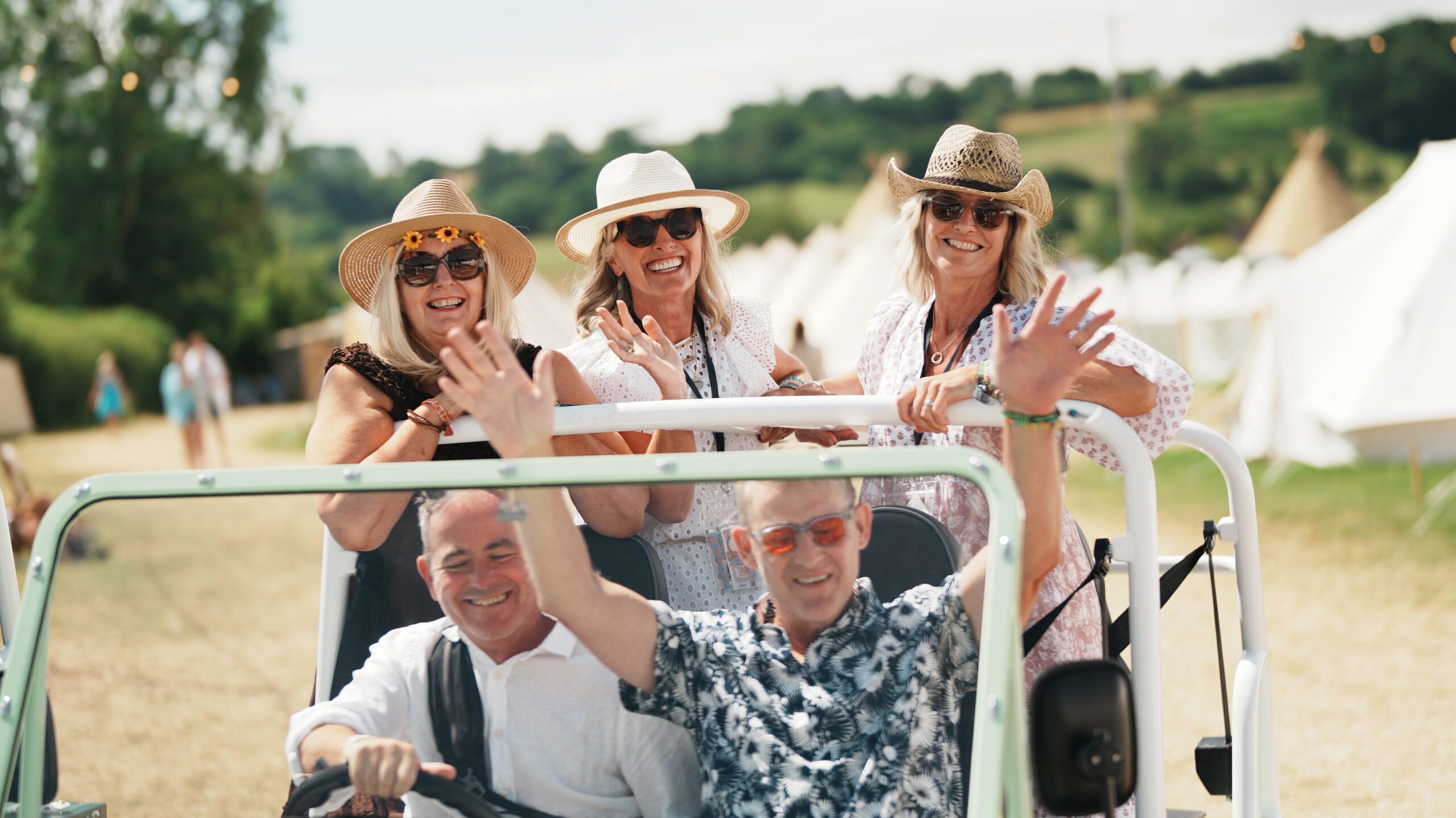 Guests in sun hats waving from a buggy as it passes the bell tents